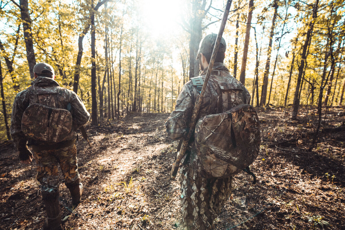 A hunter stands with his gun in the middle of the forest. He has full camouflage on and has a dog next to his leg.